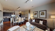 This single-floor residence offers a thoughtfully designed open-concept layout connecting the living, dining, and kitchen areas. The living room features warm hardwood-style flooring, track lighting, neutral-toned walls, and generous wall space showcasing framed artwork. A glass coffee table and ample seating create an inviting atmosphere, while large windows allow natural light to fill the space.

The kitchen is appointed with dark espresso cabinetry extending to the ceiling, granite countertops, a tile backsplash, and a full suite of stainless steel appliances. A kitchen island with an undermount sink provides additional prep space and separates the kitchen from the dining area, which comfortably accommodates a full dining set.

Both bedrooms feature plush carpeting, bold accent walls in muted teal tones, and large windows offering city views. The primary suite includes a spacious walk-in closet with built-in shelving and a private en-suite bathroom featuring a dual-sink vanity with dark cabinetry, a soaking tub, and tile flooring. The second bathroom is similarly well-equipped, maintaining a cohesive, polished aesthetic throughout the residence.