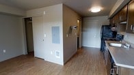 This single-floor unit offers a functional and straightforward layout designed for efficient use of space. The interior is finished with light hardwood-style flooring throughout, lending warmth and continuity to the open area. Neutral taupe and warm gray wall tones create a calm, understated ambiance that complements the clean lines of the space.

A central dividing wall serves as a practical structural element, housing an electrical panel and a wall-mounted accessory holder. To one side of this partition, a sliding-door closet provides generous storage space, while a floor-level vent ensures consistent climate control throughout the unit.

The entryway is clearly defined by a solid, white-paneled front door with a standard deadbolt lock, providing a secure and welcoming point of entry. Flush-mount ceiling lighting offers bright, even illumination across the main area. A glimpse of what appears to be a kitchenette or secondary space is visible beyond the central wall, suggesting a thoughtfully segmented floor plan.

Overall, the space presents a clean, move-in-ready interior with practical features, durable finishes, and a neutral palette that lends itself well to a variety of furnishing styles and personal preferences.