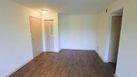 This single-floor space presents a straightforward and functional layout designed for comfortable everyday living. The main living area immediately draws the eye with its rich, dark hardwood-style flooring that runs throughout, providing a warm contrast against the clean, neutral white walls. The space feels open and uncluttered, offering a blank canvas that lends itself well to a variety of furniture arrangements and personal styles. A flush-mount ceiling light provides consistent overhead illumination throughout the room. Practicality is built into the design with a louvered bi-fold closet door near the entryway, offering convenient storage space. The entry door is solid and well-positioned, creating a clear and accessible threshold into the living area. White baseboards run along the perimeter of the room, adding a clean, finished look to the flooring transition. A ceiling vent indicates a central air and heating system for year-round climate control. The overall ambiance is bright, clean, and move-in ready, with a minimalist character that makes it easy to envision and personalize the space to suit individual needs.