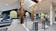 Located in Mill Creek, WA, this two-floor fitness facility offers a thoughtfully designed environment for a comprehensive workout experience. The upper level serves as the main fitness floor, characterized by soaring vaulted ceilings, large arched and rectangular windows that flood the space with natural light, and striking wood columns that add a warm, architectural contrast to the crisp white walls. A sculptural sputnik chandelier anchors the central open area, which overlooks the lower level via a sleek metal railing staircase. This floor houses a well-equipped cardio zone with treadmills and ellipticals, a dedicated strength training area with free weights, cable machines, and weight racks, and a rowing and cycling section — all framed by views of the surrounding Pacific Northwest greenery. A motivational neon sign and bold accent walls reinforce an energizing atmosphere throughout. The lower level features light hardwood flooring across two versatile open studios, ideal for yoga, stretching, or group classes. One studio is highlighted by a vivid full-wall sunset mural and wall-mounted display screen, while the other offers direct access to an outdoor patio through French doors. Together, the two floors create a well-rounded, amenity-rich fitness destination.