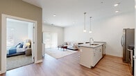 Located at 1866 E 93rd St in Cleveland, Ohio, this modern single-floor unit offers a thoughtfully designed open-concept layout that seamlessly connects the kitchen and living areas. The kitchen is a standout feature, showcasing granite countertops, crisp white cabinetry with brushed gold hardware, a large center island with a built-in sink, stainless steel appliances including a refrigerator, electric range, over-the-range microwave, and dishwasher, and stylish pendant lighting overhead. Light wood-look flooring runs throughout the main living areas, enhancing the bright and airy atmosphere. The living room is generously sized and bathed in natural light from a full-height sliding glass door dressed with vertical blinds, offering a view of the surrounding neighborhood. The bedroom features plush carpeting, a large window with an outdoor view, and ample space for furnishings. The bathroom is clean and contemporary, finished with large-format tile flooring, a white vanity, and brushed gold fixtures. A dedicated laundry area accommodates a full stacked washer and dryer unit with additional shelving. The overall design is modern and neutral, offering a comfortable and functional living environment.