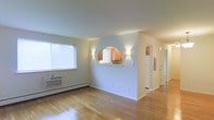 This single-floor residence is defined by its clean, open layout and warm, functional design. The living area features light-toned hardwood floors that extend throughout, creating a cohesive and inviting atmosphere. A large window fitted with horizontal blinds allows natural light to fill the space, while a wall-mounted HVAC unit and baseboard heating ensure year-round comfort.

A standout architectural detail is the arched pass-through opening in the dividing wall, which connects the living area to the kitchen. Flanked by wall sconces that cast a warm glow, this curved archway adds a distinctive design element while maintaining an open, airy feel between the two spaces. Through the pass-through, honey-toned wood cabinetry is visible, offering a warm contrast to the neutral white walls.

A hallway visible from the main living area leads further into the unit, passing what appears to be a white refrigerator and additional storage or utility areas. The overall color palette—soft whites, warm wood tones, and natural light—creates a bright and welcoming environment throughout the space.