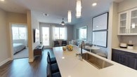 This contemporary single-floor residence showcases a thoughtfully designed open-concept layout that seamlessly integrates the kitchen, dining, and living areas. The kitchen is a standout feature, equipped with stainless steel appliances including a gas range, built-in microwave, and French door refrigerator. Two-toned cabinetry pairs dark wood lower cabinets with crisp white uppers, complemented by a tile backsplash and a generous island with a white quartz countertop and undermount stainless steel sink. Pendant lighting adds a refined touch overhead. The living area is warm and inviting, featuring dark hardwood-style flooring, a ceiling fan, and access to an outdoor balcony through glass-paneled doors, allowing ample natural light to fill the space. A large mirror and curated wall art enhance the room's airy ambiance. The bedroom offers plush carpeting, a spacious walk-in closet, and a bright window with sheer curtains that frame views of the surrounding neighborhood. A rich wood dresser adds warmth to the room. The bathroom is elegantly finished with wood-look tile flooring, a dual-sink vanity with dark cabinetry, a soaking tub with tiled surround, and a separate glass-enclosed shower, creating a serene, spa-like retreat.
