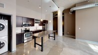 This single-floor unit showcases an industrial-modern aesthetic that blends raw architectural character with contemporary finishes. The open-concept kitchen serves as a central highlight, featuring dark espresso cabinetry, a white subway tile backsplash, stainless steel appliances including a refrigerator, electric range, microwave, and dishwasher, and a large kitchen island with a sleek quartz-style countertop supported by bold metal legs. Pendant lighting hangs from soaring ceilings, emphasizing the generous vertical space. Exposed ductwork, structural columns, and a concrete ceiling reinforce the building's industrial origins, adding a loft-like character throughout. The in-unit stacked washer and dryer are conveniently located adjacent to the kitchen. The living areas feature polished concrete-style tile flooring that reflects natural light, while the bedroom is carpeted and benefits from large industrial-style windows with grid detailing that frame urban views. The full bathroom is clean and functional, featuring a dark vanity with a white countertop and integrated sink, a tub-shower combination, and chrome fixtures throughout, maintaining the unit's consistent modern palette.