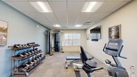 Located at 88 Oldfield Dr in Charlottesville, Virginia, this well-equipped exercise room offers a focused and functional fitness environment. The space is thoughtfully organized to accommodate a diverse range of workout routines within a single-floor layout. Underfoot, neutral-toned carpet tile flooring covers the entire room, while a drop panel ceiling with recessed fluorescent lighting ensures consistent, bright illumination throughout. A pair of windows along one wall admits natural light and provides views of the surrounding exterior, lending the room an open, airy feel. The cardio zone features multiple Matrix-brand machines, including ellipticals and a stationary bike, positioned near the windows to make the most of the natural light. A full-frame Matrix cable machine anchors the strength-training area, offering versatile resistance options. A three-tier dumbbell rack lines the accent wall, which is finished in a calming muted blue, and holds a complete set of free weights for targeted strength work. An adjustable workout bench occupies the center of the room for flexibility in exercise routines. A wall-mounted flat-screen television adds an entertainment option for users during workouts. Framed artwork on the walls and a clean, organized layout contribute to a pleasant and motivating atmosphere. A private restroom is accessible directly from the exercise room, adding convenience for users.
