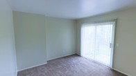 This two-story space features a clean, understated design with neutral tones throughout. The pictured room showcases light beige carpeting paired with soft, cream-colored walls, creating a calm and functional environment. A large sliding glass door fitted with vertical blinds serves as a prominent architectural feature, allowing abundant natural light to fill the room while providing access to an outdoor area. The blinds offer adjustable privacy and light control, making the space versatile throughout the day. The room's layout is open and unobstructed, providing flexibility for various furniture arrangements. Smooth white ceilings contribute to the airy feel of the room. A floor-level air vent is visible near the sliding door, indicating a built-in climate control system. The walls are clean with minimal detailing, and white baseboards provide a crisp border between the walls and carpet. Spanning two floors, this property offers distinct living zones, allowing for separation between common areas and private spaces. Overall, the space maintains a practical, move-in-ready condition with a neutral palette that accommodates a wide range of interior styles.