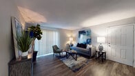 This single-story unit offers a well-organized layout with distinct living, sleeping, and functional spaces. The living room serves as the centerpiece, featuring rich wood-look flooring, a large sliding glass door fitted with vertical blinds that invite natural light, and neutral walls that complement bold accent artwork. The open, airy feel is enhanced by smooth ceilings and warm ambient lighting throughout.

The galley-style kitchen is efficiently designed with dark cabinetry, stone-look countertops, a tile backsplash, and black appliances including a refrigerator and range. Wood-look flooring continues here, maintaining visual consistency with the main living area.

The primary bedroom is a standout space, featuring a dramatic navy accent wall, plush carpeting, a ceiling fan with integrated lighting, and windows dressed with curtain panels for light control. A second bedroom offers a similarly comfortable atmosphere with neutral tones, carpeted floors, a ceiling fan, and a window allowing natural light.

The bathroom is clean and functional, featuring a full tub-shower combination with tile surround, safety grab bars, wood-look flooring, and dark vanity cabinetry with stone-look countertops. Overall, the unit presents a cohesive, modern aesthetic with a navy and grey color palette carried throughout.