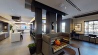 Located at 6600 Main St in Miami Lakes, Florida, this single-floor amenity space offers a thoughtfully designed environment that blends social, recreational, and functional areas into one cohesive layout. The interior showcases a contemporary aesthetic defined by dark wood-paneled columns, large-format tile flooring, coffered and tray ceilings with recessed and ambient lighting, and an open floor plan that connects each zone fluidly.

The lounge area features plush, channel-tufted seating in metallic and cool-toned upholstery, arranged around the signature dark columns that anchor the space architecturally. Floor-to-ceiling windows and glass-panel doors flood the interior with natural light and offer views of the lush, tree-lined surroundings outside.

A fully equipped bar and kitchen station includes sleek quartz countertops, bar seating, dark wood cabinetry, a mosaic tile backsplash, and mounted television — ideal for social gatherings. Adjacent to this is a dining area with a live-edge wood table, sculptural pendant lighting, and an interior window that connects visually to a private conference or meeting room beyond.

The game room provides a pool table, foosball table, wall-mounted displays, and curated artwork, all set beneath a dramatic layered ceiling. A dedicated workstation nook with a counter-height desk and garden views rounds out this versatile, amenity-rich space.