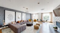 This thoughtfully designed single-floor amenity space offers a variety of distinct zones tailored for socializing, relaxing, and gathering. The main lounge area makes an immediate impression with its textured dark accent wall adorned with large-scale botanical and geometric artwork, serving as a dramatic backdrop to a generously sized tufted gray sectional sofa accented with colorful throw pillows. A sculptural black organic-form coffee table anchors the seating arrangement, complemented by woven rattan poufs and a soft blue area rug underfoot. A sleek brass-and-black sputnik-style chandelier provides warm overhead illumination throughout the lounge.

Adjacent to the main seating area, a dining and café nook features intimate round tables paired with vibrant mustard and emerald green velvet chairs, bathed in natural light from floor-to-ceiling windows dressed in sheer linen drapes that offer views of the surrounding greenery.

A separate multi-purpose room features a striking wood-slat ceiling, a large white island with integrated bench seating, warm wood cabinetry, and an industrial-style overhead garage door that opens to an outdoor-facing window wall, creating a seamlessly connected indoor-outdoor feel. Light hardwood flooring flows throughout, unifying the space with a clean, contemporary aesthetic.
