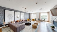 This thoughtfully designed single-floor amenity space offers a variety of distinct zones tailored for socializing, relaxing, and gathering. The main lounge area makes an immediate impression with its textured dark accent wall adorned with large-scale botanical and geometric artwork, serving as a dramatic backdrop to a generously sized tufted gray sectional sofa accented with colorful throw pillows. A sculptural black organic-form coffee table anchors the seating arrangement, complemented by woven rattan poufs and a soft blue area rug underfoot. A sleek brass-and-black sputnik-style chandelier provides warm overhead illumination throughout the lounge.

Adjacent to the main seating area, a dining and café nook features intimate round tables paired with vibrant mustard and emerald green velvet chairs, bathed in natural light from floor-to-ceiling windows dressed in sheer linen drapes that offer views of the surrounding greenery.

A separate multi-purpose room features a striking wood-slat ceiling, a large white island with integrated bench seating, warm wood cabinetry, and an industrial-style overhead garage door that opens to an outdoor-facing window wall, creating a seamlessly connected indoor-outdoor feel. Light hardwood flooring flows throughout, unifying the space with a clean, contemporary aesthetic.