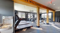 This single-floor fitness center is designed with a bold, energetic aesthetic that reflects its purpose. The interior is defined by dark charcoal walls accented by striking yellow-orange structural columns and overhead beams, one of which displays the motivational phrase, "This Place Will Change You If You Let It," reinforcing the facility's active atmosphere.

The space is divided into distinct functional zones. The cardio area is lined with multiple treadmills, elliptical trainers, stair climbers, and a stationary bike, all arranged along walls lined with large windows that allow generous natural light and views of the surrounding exterior. Ceiling fans are mounted throughout to support air circulation during workouts.

A dedicated strength training zone features a dumbbell rack with a full range of weights, adjustable weight benches, and a cable machine, with a large wall mirror enhancing visibility and the sense of space. The flooring throughout consists of patterned modular carpet tiles in grey and tan tones.

The entrance area includes a secured glass door entry with a controlled access system, offering a polished and functional welcome point. Mounted televisions are also present, adding an entertainment element to the workout environment.