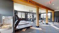 This single-floor fitness center is designed with a bold, energetic aesthetic that reflects its purpose. The interior is defined by dark charcoal walls accented by striking yellow-orange structural columns and overhead beams, one of which displays the motivational phrase, "This Place Will Change You If You Let It," reinforcing the facility's active atmosphere.

The space is divided into distinct functional zones. The cardio area is lined with multiple treadmills, elliptical trainers, stair climbers, and a stationary bike, all arranged along walls lined with large windows that allow generous natural light and views of the surrounding exterior. Ceiling fans are mounted throughout to support air circulation during workouts.

A dedicated strength training zone features a dumbbell rack with a full range of weights, adjustable weight benches, and a cable machine, with a large wall mirror enhancing visibility and the sense of space. The flooring throughout consists of patterned modular carpet tiles in grey and tan tones.

The entrance area includes a secured glass door entry with a controlled access system, offering a polished and functional welcome point. Mounted televisions are also present, adding an entertainment element to the workout environment.