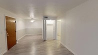 This single-floor residence is designed with a straightforward, functional layout that maximizes the available living space. The main living area greets occupants through a warm wood-toned entry door and features light gray luxury vinyl plank flooring, neutral white walls with subtle two-tone detailing, and a ceiling fan that aids in air circulation. A bi-fold closet door near the living area provides convenient built-in storage.

The kitchen is efficiently arranged in a galley-style configuration, equipped with a coil-burner electric range, a built-in range hood, a refrigerator, and a single-basin stainless steel sink. Light wood-toned upper cabinetry lines the walls, offering ample storage, while white laminate countertops provide a clean, practical workspace. An open pass-through connects the kitchen directly to the main living area.

The bedroom is a well-proportioned, carpeted room featuring a window with blinds that allows natural light to filter in, contributing to a comfortable atmosphere.

The bathroom is finished with small mosaic floor tiles, a vanity with under-sink cabinet storage, a mirror, and a shower-tub combination enclosed by a curtain. Overall, the space presents a clean, neutral palette throughout.