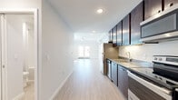 This single-floor unit offers a thoughtfully designed layout encompassing a living room, kitchen, and bathroom. Light wood-look flooring runs throughout, lending a clean and cohesive aesthetic to the entire space.

The galley-style kitchen is a standout feature, outfitted with rich dark wood cabinetry that extends floor to ceiling, providing ample storage. Stainless steel appliances — including a refrigerator, range, built-in microwave, and dishwasher — complement the sleek grey countertops. Under-cabinet lighting adds both functionality and ambiance to the workspace.

The living room is bright and open, benefiting from natural light that streams through a set of French doors, which offer outdoor access and views beyond. A wall-mounted mini-split HVAC unit ensures efficient climate control. Recessed ceiling lighting and a ceiling fan further enhance the comfort and functionality of this area.

A separate room with a built-in closet shelving system and a tall window fills the space with natural light, offering versatile use as a bedroom or home office.

The bathroom features a full bathtub and shower combination with brushed nickel fixtures set against clean white walls, maintaining the modern, minimalist character found throughout the unit. The overall design emphasizes simplicity, functionality, and a bright, airy atmosphere.