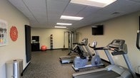 This fitness center is a dedicated, single-floor exercise facility designed to support a well-rounded workout routine. The room is outfitted with a selection of cardio equipment, including a treadmill, an elliptical trainer, and a stationary recumbent bike, all arranged to allow comfortable use and easy movement between machines. A multi-functional cable resistance station anchors the strength training area, complemented by additional accessories such as a stability ball and a rack of handheld weights, catering to both resistance and flexibility training. A wall-mounted flat-screen television is positioned for convenient viewing during workouts. The flooring throughout the space consists of durable, shock-absorbing rubber matting, which is practical for exercise use and easy maintenance. A drop ceiling with recessed fluorescent panel lighting provides consistent, even illumination across the room. A storage cabinet near the entrance offers space for towels or other fitness supplies. The neutral-toned walls and functional layout create a clean, focused environment well-suited for daily fitness activity.