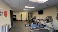 This fitness center is a dedicated, single-floor exercise facility designed to support a well-rounded workout routine. The room is outfitted with a selection of cardio equipment, including a treadmill, an elliptical trainer, and a stationary recumbent bike, all arranged to allow comfortable use and easy movement between machines. A multi-functional cable resistance station anchors the strength training area, complemented by additional accessories such as a stability ball and a rack of handheld weights, catering to both resistance and flexibility training. A wall-mounted flat-screen television is positioned for convenient viewing during workouts. The flooring throughout the space consists of durable, shock-absorbing rubber matting, which is practical for exercise use and easy maintenance. A drop ceiling with recessed fluorescent panel lighting provides consistent, even illumination across the room. A storage cabinet near the entrance offers space for towels or other fitness supplies. The neutral-toned walls and functional layout create a clean, focused environment well-suited for daily fitness activity.