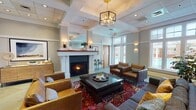 Located at 810 Catalpa Ct in Charlottesville, Virginia, this single-floor community space is thoughtfully designed to offer both comfort and functionality. The centerpiece of the space is a refined lounge area anchored by a classic white-surround fireplace, which creates a warm and inviting atmosphere. The lounge is furnished with leather armchairs and a plush sectional sofa arranged around a dark wood coffee table, all set atop a vibrant traditional area rug. A stylish geometric brass pendant chandelier hangs from a coffered ceiling, adding an upscale touch to the room. Large windows along the perimeter flood the space with natural light and offer views of the surrounding outdoor greenery. An open shelving unit serves as a beverage and accessory station, adding practical convenience to the lounge. Adjacent to the main lounge is a dedicated dining nook featuring a round wooden table surrounded by cane-back chairs, lit by tall windows that bring in generous daylight and botanical wall art that adds a natural, curated aesthetic. The kitchen is equipped with stainless steel appliances, white cabinetry, and light stone-look countertops, providing a clean and functional space for food preparation. Throughout, the neutral gray and white color palette, tile flooring, and refined architectural details create a cohesive and polished environment.