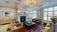 Located at 810 Catalpa Ct in Charlottesville, Virginia, this single-floor community space is thoughtfully designed to offer both comfort and functionality. The centerpiece of the space is a refined lounge area anchored by a classic white-surround fireplace, which creates a warm and inviting atmosphere. The lounge is furnished with leather armchairs and a plush sectional sofa arranged around a dark wood coffee table, all set atop a vibrant traditional area rug. A stylish geometric brass pendant chandelier hangs from a coffered ceiling, adding an upscale touch to the room. Large windows along the perimeter flood the space with natural light and offer views of the surrounding outdoor greenery. An open shelving unit serves as a beverage and accessory station, adding practical convenience to the lounge. Adjacent to the main lounge is a dedicated dining nook featuring a round wooden table surrounded by cane-back chairs, lit by tall windows that bring in generous daylight and botanical wall art that adds a natural, curated aesthetic. The kitchen is equipped with stainless steel appliances, white cabinetry, and light stone-look countertops, providing a clean and functional space for food preparation. Throughout, the neutral gray and white color palette, tile flooring, and refined architectural details create a cohesive and polished environment.