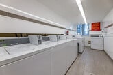 This shared laundry facility is a compact yet efficiently organized space designed to accommodate multiple users simultaneously. The room is laid out in a long, narrow corridor-style configuration, maximizing the use of available floor space along both walls. White large-format ceramic tiles line the walls with a subtle tan accent band running horizontally at mid-height, contributing to the clean, modern aesthetic. The light-toned tile flooring complements the overall bright and hygienic atmosphere.

A row of top-loading washing machines lines one side of the room, paired with front-loading dryers equipped with digital controls on the opposite side. A stainless steel folding table provides a convenient surface for handling laundry. Overhead, continuous fluorescent strip lighting ensures consistent, even illumination throughout the entire space. A window at the far end of the room allows natural light to supplement the artificial lighting.

Safety is addressed with a mounted fire extinguisher and posted signage, including laundry room rules and notices displayed prominently on the walls. A secure entry door with a glass panel provides controlled access to the facility. The space is well-maintained, presenting a tidy and functional environment suited for regular communal use.
