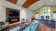 Located at 916 Sugar Gap Rd in Wake Forest, this single-story community amenity building offers a thoughtfully designed environment suited for both relaxation and active living. The main clubroom is an eye-catching space defined by soaring vaulted ceilings, bold chartreuse accent walls, and an expansive wall of arched windows that flood the interior with natural light and offer views of the surrounding greenery. Rich hardwood flooring runs throughout, complemented by vibrant patterned area rugs that add color and warmth. A striking focal wall features a wall-mounted television above a sleek linear electric fireplace framed in warm wood paneling, creating an inviting gathering area with plush seating. A decorative wave-textured panel divides the space, providing a sense of intimacy within the open layout. A dedicated workstation area with computer access offers a quiet nook for productivity. Adjacent to the clubroom, a casual café-style lounge provides high-top seating and a coffee station, with direct access to the outdoor patio through glass doors. The kitchenette is appointed with modern cabinetry, a mosaic tile backsplash in shades of blue, and ample counter space. The fitness center is well-equipped with a comprehensive selection of cardio machines, strength-training equipment, free weights, and a wall-mounted television, all set on durable multi-tone flooring beneath ceiling fans and ample lighting.