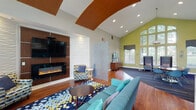 Located at 916 Sugar Gap Rd in Wake Forest, this single-story community amenity building offers a thoughtfully designed environment suited for both relaxation and active living. The main clubroom is an eye-catching space defined by soaring vaulted ceilings, bold chartreuse accent walls, and an expansive wall of arched windows that flood the interior with natural light and offer views of the surrounding greenery. Rich hardwood flooring runs throughout, complemented by vibrant patterned area rugs that add color and warmth. A striking focal wall features a wall-mounted television above a sleek linear electric fireplace framed in warm wood paneling, creating an inviting gathering area with plush seating. A decorative wave-textured panel divides the space, providing a sense of intimacy within the open layout. A dedicated workstation area with computer access offers a quiet nook for productivity. Adjacent to the clubroom, a casual café-style lounge provides high-top seating and a coffee station, with direct access to the outdoor patio through glass doors. The kitchenette is appointed with modern cabinetry, a mosaic tile backsplash in shades of blue, and ample counter space. The fitness center is well-equipped with a comprehensive selection of cardio machines, strength-training equipment, free weights, and a wall-mounted television, all set on durable multi-tone flooring beneath ceiling fans and ample lighting.