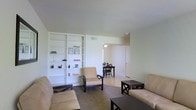 This single-floor space presents a straightforward and functional layout, with an open living area serving as its central hub. The living room is furnished with a sofa and accent chairs in neutral tones, complemented by a dark-framed coffee table and side table, creating a modest yet practical seating arrangement. A standout architectural feature is the large built-in white shelving unit along one wall, offering ample display and storage space. The shelves are dressed with small decorative items, framed pieces, and greenery, adding a personal touch to the room. Light-colored walls and a smooth ceiling contribute to a clean, airy feel throughout. The flooring appears to be a dark, low-pile carpet, grounding the space with a warm texture. An open passageway connects the living area to a dining space, where a dark wooden table and chairs are visible, suggesting a seamless flow between the two areas. A door beyond the dining space hints at additional rooms. Natural light filters in through a window in the dining area, brightening the overall environment. The space maintains a calm, understated ambiance suited for everyday living.