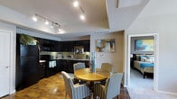 This single-floor residence showcases an open-concept layout that seamlessly connects the kitchen and dining area, creating a cohesive and functional living space. The kitchen is outfitted with rich, dark wood cabinetry that extends to the upper walls, providing generous storage while lending a bold, contemporary aesthetic. Black appliances, including a range and built-in microwave, complement the cabinetry, while the countertops offer ample workspace. A sink positioned along the lower cabinetry run adds to the kitchen's practical design.

The dining area sits adjacent to the kitchen, centered around a warm, round wooden table surrounded by striped upholstered chairs that add a classic touch. Vaulted ceilings overhead enhance the sense of openness, while a track lighting system illuminates both the kitchen and dining spaces effectively. The flooring transitions from warm hardwood in the main living area to carpet visible in the adjoining bedroom, which can be seen through a doorway at the far end of the space. Neutral wall tones and tasteful artwork contribute to the overall inviting and well-appointed ambiance.
