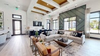 This single-floor communal lounge space makes a striking architectural statement through its dramatic double-height ceilings and carefully curated design elements. The main gathering area is anchored by a coffered ceiling finished with warm gold-toned inlays and illuminated by elegant brass pendant lights, creating a refined focal point overhead. Light-toned hardwood flooring flows throughout, unifying the open-plan layout and complementing the bright, airy atmosphere.

Two distinct accent walls define the space: a textured, crackle-finish white wall housing a wall-mounted television and a bold geometric green paneled wall that rises dramatically to the full ceiling height. Large black-framed windows and glass partition walls flood the interior with natural light while offering views of the surrounding outdoor landscape, including palm trees and neighboring structures.

The seating areas are thoughtfully arranged with neutral sectional sofas, caramel leather accent chairs with gold frames, and a mix of round and rectangular coffee tables, creating comfortable zones for socializing or relaxing. A separate lounge nook with swivel chairs and additional wall-mounted displays provides a quieter retreat. The overall ambiance is modern and upscale, balancing bold design choices with inviting comfort.
