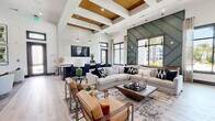 This single-floor communal lounge space makes a striking architectural statement through its dramatic double-height ceilings and carefully curated design elements. The main gathering area is anchored by a coffered ceiling finished with warm gold-toned inlays and illuminated by elegant brass pendant lights, creating a refined focal point overhead. Light-toned hardwood flooring flows throughout, unifying the open-plan layout and complementing the bright, airy atmosphere.

Two distinct accent walls define the space: a textured, crackle-finish white wall housing a wall-mounted television and a bold geometric green paneled wall that rises dramatically to the full ceiling height. Large black-framed windows and glass partition walls flood the interior with natural light while offering views of the surrounding outdoor landscape, including palm trees and neighboring structures.

The seating areas are thoughtfully arranged with neutral sectional sofas, caramel leather accent chairs with gold frames, and a mix of round and rectangular coffee tables, creating comfortable zones for socializing or relaxing. A separate lounge nook with swivel chairs and additional wall-mounted displays provides a quieter retreat. The overall ambiance is modern and upscale, balancing bold design choices with inviting comfort.