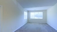 This single-floor space features a spacious main room finished with wall-to-wall carpeting in a neutral tone, contributing to a clean and understated aesthetic. The room is characterized by its simplicity and openness, with smooth, light-colored walls that complement the overall bright atmosphere. Two windows are positioned to maximize natural light — a single window on the side wall and a larger multi-pane window on the facing wall — both allowing views of the surrounding outdoor greenery. The windows are fitted with horizontal blinds, offering flexible light control. White baseboards line the perimeter of the room, providing a crisp contrast against the neutral carpet and walls. A visible ceiling vent suggests a central climate control system is in place. The absence of furniture allows the full scale of the room to be appreciated, revealing a generous, versatile layout well-suited for a variety of uses. The overall ambiance is calm and neutral, providing a blank canvas that can be easily adapted to different interior styles and functional needs.