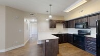 Located at 1835 N Berra Blvd in Tooele, Utah, this well-appointed 2-bedroom, 2-bathroom single-floor residence offers a thoughtfully designed layout across a comfortable footprint. The open-concept kitchen and living area serves as the heart of the home, featuring rich dark espresso cabinetry with brushed nickel hardware, crisp white countertops, a central island with pendant lighting, and a full suite of black appliances including an electric range, built-in microwave, dishwasher, and refrigerator. Wood-style flooring runs throughout the kitchen and into the living room, which is bathed in natural light from a sliding glass door leading to an outdoor patio. A ceiling fan adds both style and functionality to the living area. A hallway connects the main living space to the private quarters, where two carpeted bedrooms offer a warm, quiet retreat. The spacious primary bedroom includes its own walk-in closet with built-in shelving. Both bathrooms are finished with tile flooring and include tub/shower combinations. A dedicated utility room provides added convenience. Neutral warm tones throughout create a cohesive and inviting atmosphere.