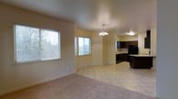 This single-floor residence offers an open-concept layout that seamlessly connects the living, dining, and kitchen areas into one cohesive and functional space. The living area is finished with soft, neutral-toned carpet, providing a comfortable and inviting atmosphere, while the dining and kitchen zones transition to durable tile flooring. Multiple windows fitted with horizontal blinds allow generous natural light to fill the space, offering views of lush greenery outside. A decorative chandelier hangs above the dining area, adding a refined touch to the overall design. The kitchen is well-appointed with rich, dark espresso cabinetry that creates a striking contrast against the light-colored countertops and tile backsplash. Black appliances, including a full-size refrigerator, complement the kitchen's contemporary aesthetic. Under dedicated overhead lighting, the kitchen provides ample workspace and storage. The warm, cream-toned walls throughout the space contribute to a bright and airy ambiance. The open flow between rooms makes the layout feel expansive and versatile, suitable for both everyday living and entertaining.