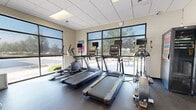 This fitness center offers a well-rounded workout environment housed within a single, open-plan room designed to accommodate a wide variety of exercise needs. The space is lined with large windows along multiple walls, flooding the interior with abundant natural light and providing views of the surrounding outdoor area. Roller shades allow for adjustable light control throughout the day.

The cardio section features multiple treadmills equipped with individual monitors, along with elliptical trainers and a stair-stepper, all arranged to face the expansive windows. A row of upright and recumbent stationary bikes provides additional low-impact options. The strength training area includes a multi-station cable machine, a dedicated weight bench, and a selection of free weights, offering comprehensive resistance training options.

A large wall mirror expands the visual depth of the space and supports proper form during exercise. A wall-mounted flat-screen television is positioned for easy viewing from multiple workout stations. Workout towels and sanitizing supplies are readily available on a dedicated rack, promoting cleanliness. The durable rubber flooring throughout provides cushioning and traction. A drop ceiling with recessed lighting complements the natural light, ensuring the space remains bright and functional at all hours.