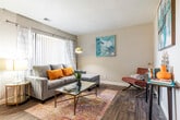 This single-floor residence offers a thoughtfully designed layout that balances functionality with contemporary style. The living room serves as the heart of the home, featuring rich dark hardwood-style flooring, a textured ceiling, and large windows dressed with sheer curtains that invite natural light. The space is anchored by a sectional sofa and a glass-top coffee table, complemented by bold abstract artwork adorning the neutral-toned walls, creating a vibrant yet cohesive atmosphere.

Adjacent to the living area, the dining space flows seamlessly from the kitchen and is illuminated by a ceiling light fixture. A round dining table with wooden chairs provides a welcoming setting for meals. The kitchen is outfitted with white cabinetry, light countertops, a gas range, and a stainless steel sink, offering a clean and practical cooking environment.

The bedroom is a serene retreat, carpeted for comfort and filled with natural light through large windows with vertical blinds. A spacious closet provides generous storage, while the neutral color palette contributes to a calm and restful ambiance. A full bathroom with white cabinetry is conveniently accessible from the main living areas. Throughout the residence, the consistent use of warm tones and curated décor creates a cohesive and inviting living environment.
