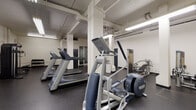 Located at 950 SW 21st Ave in Portland, Oregon, this fitness center offers a comprehensive range of exercise equipment across a single-floor layout designed for effective workouts. The space features durable rubber flooring throughout, providing a safe and practical surface for all types of training activities. Overhead fluorescent lighting ensures the entire area is well-illuminated, and exposed structural columns and utility piping give the facility an industrial character.

The cardio zone is equipped with multiple treadmills and a Precor elliptical trainer, with a wall-mounted flat-screen television positioned for use during cardio sessions. Strength training is well-supported by an Inspire functional trainer cable machine, a multi-station weight machine, and a dedicated free weights area stocked with a rack of dumbbells and an adjustable workout bench. An additional adjustable bench provides versatile options for a variety of exercises.

The layout is thoughtfully organized, with cardio equipment, strength machines, and free weight areas each occupying distinct zones within the open floor plan. Access points are provided via multiple doors, including a navy blue entry door. The overall ambiance is utilitarian and focused, offering residents a practical and well-equipped environment for maintaining an active lifestyle in the heart of Portland.