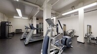 Located at 950 SW 21st Ave in Portland, Oregon, this fitness center offers a comprehensive range of exercise equipment across a single-floor layout designed for effective workouts. The space features durable rubber flooring throughout, providing a safe and practical surface for all types of training activities. Overhead fluorescent lighting ensures the entire area is well-illuminated, and exposed structural columns and utility piping give the facility an industrial character.

The cardio zone is equipped with multiple treadmills and a Precor elliptical trainer, with a wall-mounted flat-screen television positioned for use during cardio sessions. Strength training is well-supported by an Inspire functional trainer cable machine, a multi-station weight machine, and a dedicated free weights area stocked with a rack of dumbbells and an adjustable workout bench. An additional adjustable bench provides versatile options for a variety of exercises.

The layout is thoughtfully organized, with cardio equipment, strength machines, and free weight areas each occupying distinct zones within the open floor plan. Access points are provided via multiple doors, including a navy blue entry door. The overall ambiance is utilitarian and focused, offering residents a practical and well-equipped environment for maintaining an active lifestyle in the heart of Portland.