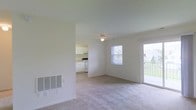 This single-floor residence offers a clean, functional layout with a light and airy feel throughout. The main living area is finished with soft wall-to-wall carpeting and neutral white walls, creating a bright and welcoming atmosphere. A ceiling fan with a light fixture is centrally positioned, providing both air circulation and ambient lighting. Natural light fills the room through a well-placed window and a glass sliding door that opens directly to an outdoor balcony, offering views of the surrounding greenery. A wall-mounted ventilation grille is integrated into the lower wall, contributing to the unit's climate control system. Adjacent to the living area, the kitchen is visible through an open pass-through layout. It features white cabinetry, white appliances including a range and what appears to be a refrigerator, and light-colored flooring, giving it a clean and practical look. The seamless flow between the living space and the kitchen enhances the overall sense of openness. The overall design is simple and uncluttered, with a neutral color palette that makes the space feel larger and easy to personalize. The combination of natural light, outdoor access, and an open plan layout makes this a versatile and comfortable living environment.