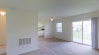 This single-floor residence offers a clean, functional layout with a light and airy feel throughout. The main living area is finished with soft wall-to-wall carpeting and neutral white walls, creating a bright and welcoming atmosphere. A ceiling fan with a light fixture is centrally positioned, providing both air circulation and ambient lighting. Natural light fills the room through a well-placed window and a glass sliding door that opens directly to an outdoor balcony, offering views of the surrounding greenery. A wall-mounted ventilation grille is integrated into the lower wall, contributing to the unit's climate control system. Adjacent to the living area, the kitchen is visible through an open pass-through layout. It features white cabinetry, white appliances including a range and what appears to be a refrigerator, and light-colored flooring, giving it a clean and practical look. The seamless flow between the living space and the kitchen enhances the overall sense of openness. The overall design is simple and uncluttered, with a neutral color palette that makes the space feel larger and easy to personalize. The combination of natural light, outdoor access, and an open plan layout makes this a versatile and comfortable living environment.