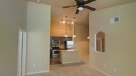 This single-story space is defined by its soaring vaulted ceilings, which lend an airy, expansive feel to the interior. The open-concept layout seamlessly connects the living area to the kitchen, creating a cohesive and functional flow throughout the space. Soft carpeting covers the living area floor, while the kitchen features hard flooring that complements the overall design.

The kitchen is well-appointed with dark espresso cabinetry, stainless steel appliances including a gas range, built-in microwave, and a full-size refrigerator. A central island with a light stone countertop provides additional workspace and serves as a natural gathering point. Pendant lights hang above the island, adding focused illumination, while a track lighting system overhead offers broader coverage across the kitchen area.

The living space benefits from a ceiling fan mounted at the peak of the vaulted ceiling, enhancing air circulation throughout. An arched architectural detail on one wall adds a decorative touch, along with a mirrored niche that reflects light and adds visual depth. The neutral warm-toned walls throughout contribute to a welcoming and cohesive ambiance.