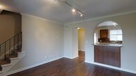 This multi-level space spans three floors, offering a thoughtfully designed interior that balances classic architectural details with modern finishes. The main living area showcases rich, dark hardwood flooring that adds warmth and depth to the space, complemented by crisp white crown molding that lines the ceilings for a refined, polished look. Track lighting provides flexible, adjustable illumination overhead, making it easy to highlight key areas of the room.

A defining architectural feature is the elegant arched passageway that connects the living area to adjacent spaces, adding a timeless character to the overall design. The kitchen is partially visible and features rich wood cabinetry, a decorative tile backsplash, and a granite or stone countertop that extends into a bar-height peninsula — ideal for casual dining or entertaining.

The neutral, light-toned walls throughout create a clean and versatile backdrop, allowing the space to feel open and airy. Hallways connect the rooms seamlessly, and the layout across three floors provides ample separation between living, sleeping, and utility areas. The combination of classic molding details, arched doorways, and quality finishes gives this space a cohesive, inviting ambiance suited for comfortable everyday living.
