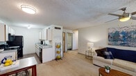 This single-story residence in Baton Rouge, Louisiana offers two bedrooms and two full bathrooms within a well-organized, functional floor plan. The open-concept living area features neutral-toned carpet and a textured ceiling, creating a bright and welcoming atmosphere. The space flows naturally from the living room toward the kitchen and bedroom corridor, maximizing the sense of openness throughout.

The galley-style kitchen is equipped with black appliances, including a range, over-the-range microwave, and refrigerator, complemented by white cabinetry with gold-tone hardware and laminate countertops. Wood-look flooring runs through the kitchen, providing a clean contrast to the carpeted living areas. Track lighting overhead ensures ample illumination for food preparation.

The primary bedroom is generously sized, featuring plush carpet, a ceiling fan with light fixture, and multiple windows that admit natural light. Grommet-style curtains provide privacy. The second bedroom is comparably comfortable, also carpeted and outfitted with a window and ceiling fan.

Both bathrooms maintain a consistent design aesthetic, featuring single-sink vanities with laminate countertops, wood-tone cabinetry, framed mirrors, and tub/shower combinations. One bathroom includes wood-look flooring, adding a modern touch. Overall, the unit presents a practical, comfortable layout suited for everyday living.