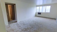 This single-floor space offers a clean and functional layout designed for comfortable everyday living. The main living area is generously sized, finished with wall-to-wall carpeting that adds warmth underfoot. Crisp white walls keep the space feeling bright and open, while a large window allows natural light to fill the room and provides a view of the surrounding outdoor environment. A wall-mounted heating and air unit ensures year-round climate control with ease. The room's open layout provides flexibility for a variety of furniture arrangements. A doorway visible from the main living area leads to an adjoining room, suggesting a practical and connected floor plan. The overall ambiance is simple, neutral, and move-in ready, offering a blank canvas for personalization. The space is well-suited for those seeking a straightforward, low-maintenance living environment with a focus on functionality and comfort.
