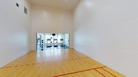 This dedicated racquetball court is a fully equipped recreational facility designed for serious play and athletic training. The court features a polished hardwood floor with clearly marked red boundary and service lines, meeting standard regulation specifications. Soaring ceilings provide ample vertical playing space, a critical element for competitive and recreational racquetball alike. The smooth, bright white walls on all sides offer a clean, consistent playing surface, minimizing visual distractions and ensuring optimal ball rebound performance. A recessed ceiling light fixture provides uniform overhead illumination throughout the court. One end of the court opens through full-height glass doors and panels, offering a seamless visual and physical connection to an adjoining fitness center. The fitness area, visible through the glass, is equipped with cardio machines including treadmills, as well as free weights, making it a convenient complement to the racquetball court. This combination of dedicated court space and adjacent gym equipment creates a comprehensive athletic environment suited for a full workout experience. The overall ambiance is clean, modern, and functional, reflecting a well-maintained facility built with athletic performance in mind.