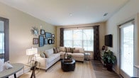 This single-floor space features a thoughtfully designed living room that serves as its central highlight. The room is finished with warm hardwood flooring and neutral beige walls that create a welcoming, versatile backdrop. A large picture window dressed with patterned draping curtains and white horizontal blinds allows abundant natural light to flow in, brightening the entire space and offering views of the greenery outside.

The living area is generously proportioned, comfortably accommodating a sectional sofa arrangement centered around a round upholstered ottoman that doubles as a coffee table. An arc floor lamp adds a stylish focal point, while a flat-screen television is positioned on the opposite wall atop a media console with storage. Recessed lighting in the smooth white ceiling supplements the natural light for evening use.

A curated gallery wall featuring an assortment of framed artwork, mirrors, and decorative accents adds personality and visual interest to the space. The overall ambiance is warm, modern, and livable, with a cohesive design aesthetic that balances comfort and style throughout.