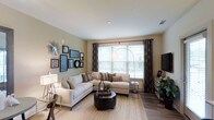 This single-floor space features a thoughtfully designed living room that serves as its central highlight. The room is finished with warm hardwood flooring and neutral beige walls that create a welcoming, versatile backdrop. A large picture window dressed with patterned draping curtains and white horizontal blinds allows abundant natural light to flow in, brightening the entire space and offering views of the greenery outside.

The living area is generously proportioned, comfortably accommodating a sectional sofa arrangement centered around a round upholstered ottoman that doubles as a coffee table. An arc floor lamp adds a stylish focal point, while a flat-screen television is positioned on the opposite wall atop a media console with storage. Recessed lighting in the smooth white ceiling supplements the natural light for evening use.

A curated gallery wall featuring an assortment of framed artwork, mirrors, and decorative accents adds personality and visual interest to the space. The overall ambiance is warm, modern, and livable, with a cohesive design aesthetic that balances comfort and style throughout.