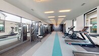 Located at 72 Milton Ave SE in Atlanta, GA, this single-floor fitness center is designed to deliver a comprehensive workout experience in a sleek, contemporary setting. The space is elongated and generously proportioned, with a clean layout that keeps cardio and strength training zones clearly defined. A series of recessed linear light fixtures runs the length of the ceiling, providing bright, even illumination throughout. Underfoot, a two-tone flooring design featuring teal accent stripes adds a dynamic visual element that guides movement through the space. The cardio zone is well-equipped with multiple treadmills, ellipticals, and a stationary bike, all positioned along expansive floor-to-ceiling windows that flood the area with natural light and offer views of the surrounding neighborhood. The strength training section includes a WaterRower, cable machines, and a multi-function power tower, complemented by mirrored walls that enhance the sense of openness. A standout feature is the dedicated amenity station at the far end of the room — a built-in countertop with cabinetry and a wine refrigerator, set against a textured teal accent wall adorned with a dramatic sunburst neon light installation. Double glass doors provide direct outdoor access, connecting the interior to the exterior grounds.