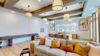 Located at 3510 Corder Dr in Wilmington, NC, this thoughtfully designed single-floor amenity space blends coastal charm with modern functionality. The main lounge area is anchored by exposed wooden ceiling beams, warm pendant lighting, and light hardwood flooring, creating an inviting and relaxed atmosphere. A generously sized sectional sofa with earth-toned accent pillows, a shuffleboard table, and a wall-mounted flat-screen TV make this area ideal for both socializing and leisure. Adjacent to the main lounge, a fully equipped kitchen features a large white quartz island, warm wood cabinetry, and an expansive window bank that floods the space with natural light and outdoor views. A secondary seating lounge offers floor-to-ceiling windows dressed with layered curtains and blinds, with mid-century modern accent chairs and intimate conversation areas. A dedicated conference and co-working room is outfitted with a central conference table, ergonomic seating, a wall-mounted display, a statement circular chandelier, and paneled gray wainscoting, providing a polished environment for professional use. Glass-paneled sliding doors provide privacy while maintaining visual connectivity. A practical hydration station with an Elkay water fountain and bottle filler, set against a decorative penny-tile mosaic wall, adds a refined functional touch throughout the facility.