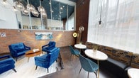 Located at 1800 S Jackson Street in Seattle's vibrant Jackson Street corridor, this single-floor commercial space combines industrial character with modern design sensibility. The interior showcases exposed brick walls in warm terra-cotta tones, contrasted by a dark ceiling with recessed lighting and a striking cluster of globe pendant lights that create an inviting, upscale ambiance. Large windows along the perimeter flood the space with natural light, while sheer curtains soften the brightness and enhance the refined atmosphere. The seating area is thoughtfully arranged with plush royal blue velvet armchairs and coordinating teal accent chairs gathered around round white bistro tables, offering a versatile layout suitable for both casual meetings and social gatherings. A bold abstract canvas painting in blues and greens adds an artistic focal point against the brick backdrop. Polished concrete floors anchor the space with an urban edge, while warm wood accents and metallic gold fixtures provide an elegant counterbalance. The combination of textures—brick, glass, metal, and fabric—creates a layered, sophisticated environment well-suited for a boutique business, lounge, or professional meeting space.