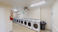 The Shrewsbury Arms laundry facility is a thoughtfully designed, single-floor communal space built to accommodate the needs of its residents with efficiency and comfort. The room is generously proportioned, featuring speckled terrazzo flooring throughout that is both durable and easy to maintain. Bright fluorescent lighting runs across the ceiling, ensuring the space is well-lit at all hours, while ceiling fans provide consistent air circulation to keep the environment comfortable during use. A wall-mounted mini-split air conditioning unit further supports a pleasant atmosphere year-round. Along one wall, a long row of modern front-loading washing machines and dryers are neatly lined up, offering substantial capacity for multiple users. A dedicated folding and sorting area is equipped with a large flat folding table, rolling laundry carts with wire baskets, and hanging racks — providing a practical and organized workflow. The warm yellow accent walls in the folding area add a welcoming touch to the otherwise neutral interior. An information and payment kiosk is conveniently located nearby. A separate open area with floor-to-ceiling glass doors leads to an outdoor greenspace, filling the room with natural light and views of surrounding landscaping, creating a bright and airy atmosphere. A fire extinguisher and waste receptacle round out the functional amenities of this well-appointed facility.