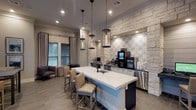 The Digital Cafe is a thoughtfully designed single-floor space that seamlessly combines a café-style atmosphere with functional workstation amenities. The interior is anchored by a striking natural stone accent wall featuring decorative patterned tile insets, lending the space a refined yet welcoming aesthetic. A central white quartz island with an undermount sink serves as the focal point, surrounded by upholstered bar-height seating and illuminated by a row of industrial-style glass pendant lights overhead.

Built-in computer workstations are recessed into arched stone niches along the back wall, each outfitted with monitors and task chairs, providing semi-private areas for focused work. Adjacent to the workstations, a fully equipped coffee bar offers a variety of beverage equipment, including a specialty coffee machine and drip brewers, complemented by stainless steel under-counter appliances.

The seating area features a mix of upholstered lounge chairs, a small occasional table, and a dining table with cushioned chairs, all positioned near large windows dressed with layered blinds and floor-length drapes. Warm wood-look flooring runs throughout, while recessed lighting and crown molding add polished finishing touches. Natural light filters through multiple windows, creating an inviting and productive environment.